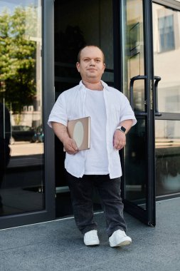 A man with inclusivity in a white shirt and black jeans holds a book and walks out of a building. clipart