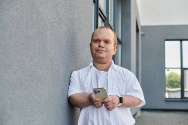 A man with inclusivity in a white shirt stands in a modern city, holding a smartphone while leaning against a wall. clipart