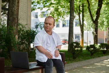 Parktaki bankta oturan ve yanında dizüstü bilgisayarı olan bir adam..