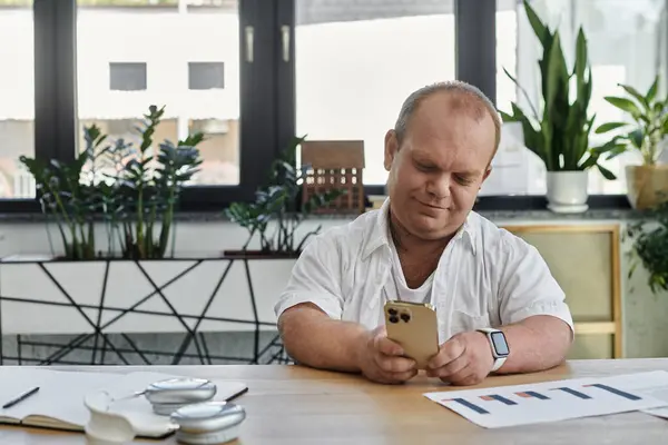 stock image A man with inclusivity sits at a desk in a modern office, checking his smartphone.