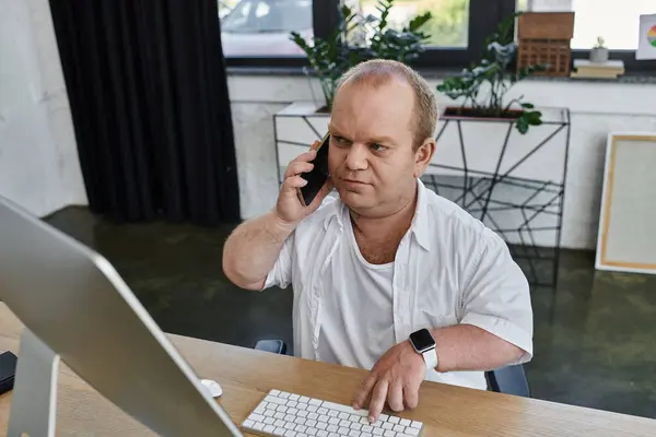 stock image A man with inclusivity in a white shirt sits at a desk in his office, using a computer and talking on a phone.