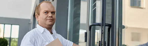 Stock image A man with inclusivity dressed in a white shirt stands by a glass door, holding a package.