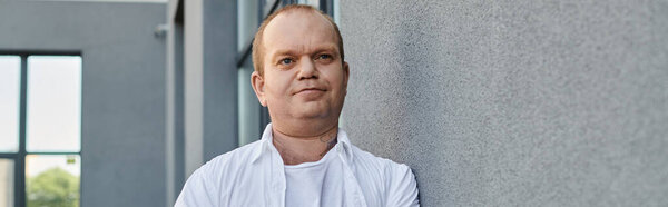 A man with inclusivity in a white shirt stands by a gray wall, looking to the side.