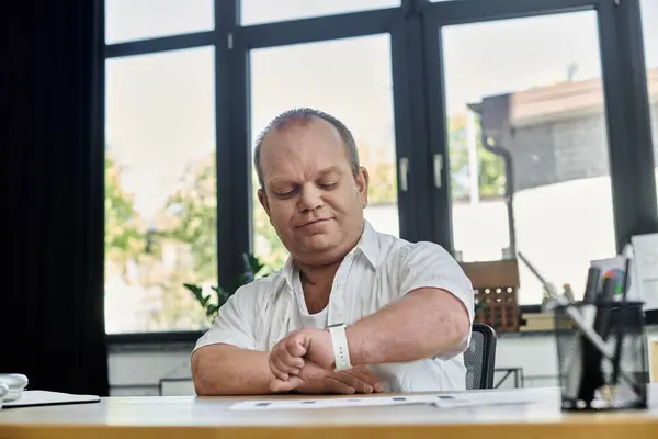 stock image A man with inclusivity in a white shirt checks the time on his wristwatch while seated at a desk in an office setting.