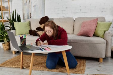 Down sendromlu küçük bir kız oturma odasındaki küçük bir masaya resim çiziyor..