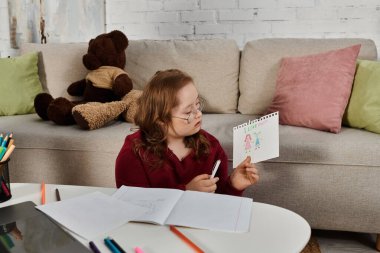 A little girl with Down syndrome shows off her drawing while sitting at a table in her home. clipart