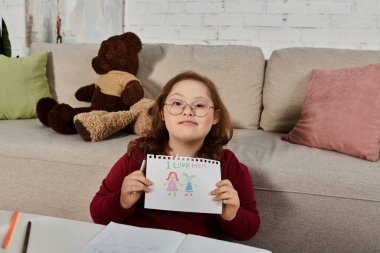 A little girl with Down syndrome sits on a couch at home, holding a drawing that says I Love Mom. clipart