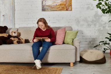 A little girl with Down syndrome sits on a couch reading a book. clipart