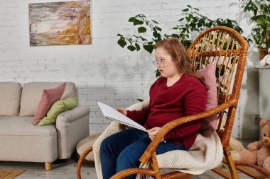 A little girl with Down syndrome sits in a rocking chair, thoughtfully reading a book. clipart