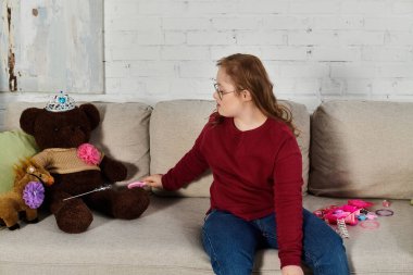 A little girl with Down syndrome plays with a teddy bear and other toys on a couch. clipart