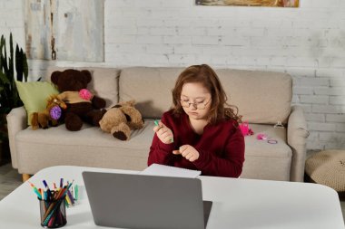 little girl with Down syndrome at desk, writing in notebook, with open laptop. clipart