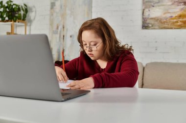 A little girl with Down syndrome sits at a table, concentrating on her work with a pencil in hand. clipart