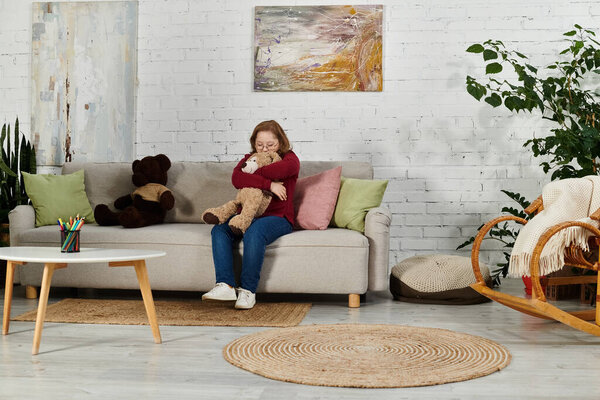 A little girl with Down syndrome sits on a couch, hugging a teddy bear, in her home.