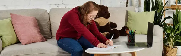 stock image Girl with Down syndrome writing on couch, notebook in hand, laptop and teddy bear nearby.