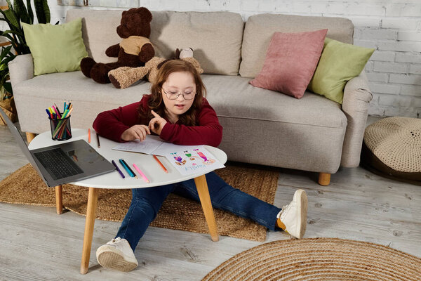 Маленькая девочка с синдромом Дауна сидит на полу в раскраске, в то время как ноутбук сидит на столе неподалеку.