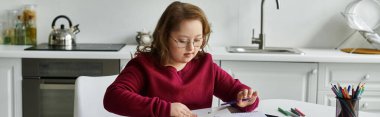 A little girl with Down syndrome sits at a table in her home, intently focusing on her drawing. clipart