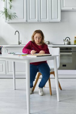 A little girl with Down syndrome sits at a kitchen table, diligently drawing or writing. clipart