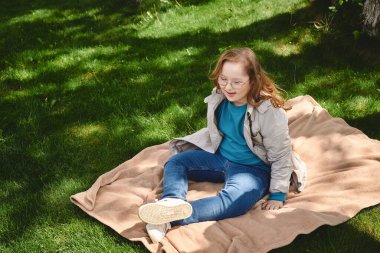 Down sendromlu küçük bir kız güneşli bir günde çimlerin üzerinde bir battaniyeye oturur..