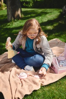 A little girl with Down syndrome enjoys a tea party with her doll in the park. clipart