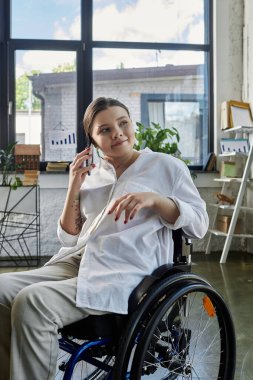 Tekerlekli sandalyedeki genç bir iş kadını modern bir ofiste çalışırken telefonunu kullanıyor..