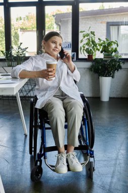 Tekerlekli sandalyedeki genç bir iş kadını modern ofis ortamında çalışıyor. Elinde kahve fincanı var ve telefonuyla konuşuyor..
