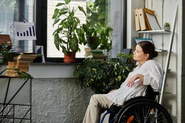 Tekerlekli sandalyedeki genç bir iş kadını modern bir ofiste çalışıyor. Doğal ışık ve bitkilerle..