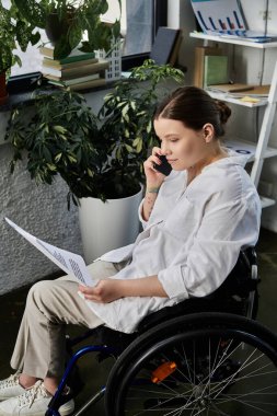 Engelli bir iş kadını modern bir ofiste tekerlekli sandalyesinde oturuyor ve bir belgeyi incelerken telefon görüşmesi yapıyor..