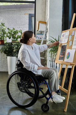 Tekerlekli sandalyedeki genç bir iş kadını modern bir ofiste oturuyor ve çizelgeleri beyaz tahtada gözden geçiriyor..
