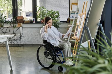 Engelli bir iş kadını modern bir ofiste tekerlekli sandalyede oturuyor, bir proje üzerinde çalışıyor ve telefonda konuşuyor..