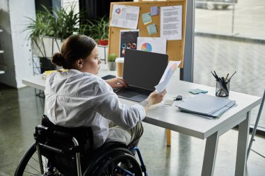 Tekerlekli sandalyedeki genç bir iş kadını, modern bir ofis masasında çalışıyor..