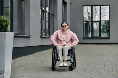 Pembe kapüşonlu genç bir kadın şehir caddesinde tekerlekli sandalyede oturuyor..