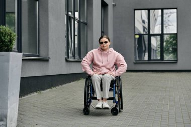 A young woman in a pink hoodie sits in a wheelchair on a city street. clipart