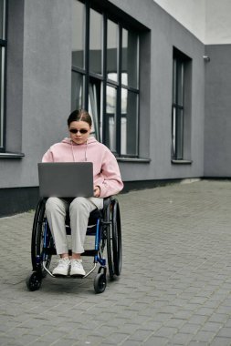 Pembe kapüşonlu genç bir kadın tekerlekli sandalyede oturuyor ve bir binanın önündeki dizüstü bilgisayarda çalışıyor..