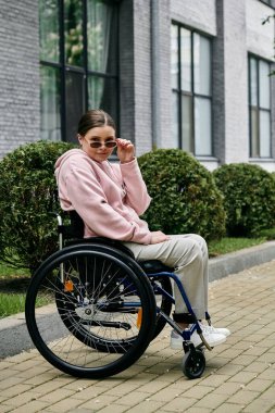 Pembe kapüşonlu genç bir kadın kaldırımda tekerlekli sandalyede oturuyor, güneş gözlüğünü kendine güvenen bir ifadeyle ayarlıyor..
