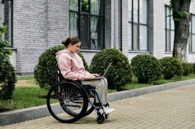 Tekerlekli sandalyedeki genç bir kadın binanın dışında kaldırımda otururken dizüstü bilgisayar kullanıyor..