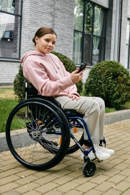 Dışarıda tekerlekli sandalyede oturan pembe kapüşonlu genç bir kadın telefonunu kullanırken gülümsüyor..