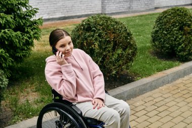 Tekerlekli sandalyede pembe kapüşonlu bir kadın oturuyor ve açık havada güneşli bir günün tadını çıkarırken telefonla konuşuyor..