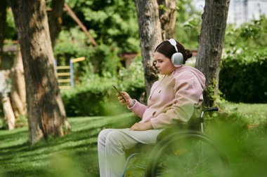 Tekerlekli sandalyedeki genç bir kadın parkta otururken telefonundan müzik dinliyor. Kulaklık ve pembe bir kapüşonlu giyiyor..