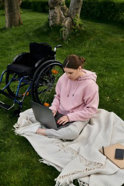 Engelli bir kadın parkta tekerlekli sandalyede oturuyor ve dizüstü bilgisayarla çalışıyor..