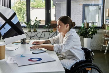 Tekerlekli sandalyedeki genç bir iş kadını modern ofis ortamında bir bilgisayarda çalışıyor..