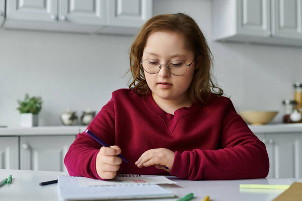 A little girl with Down syndrome sits at a table, focused on coloring with a purple pencil.