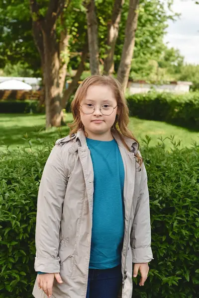 Down sendromlu küçük bir kız bahçesinde duruyor, yeşil ağaçlar ve çalılarla çevrili..