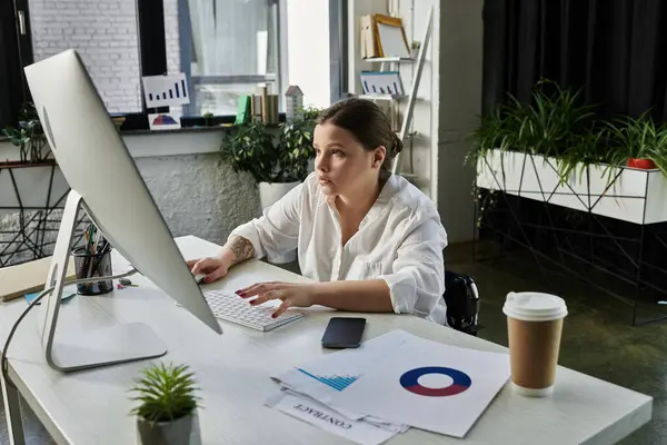 stock image A young businesswoman sits in her wheelchair working at a desk in a modern office.