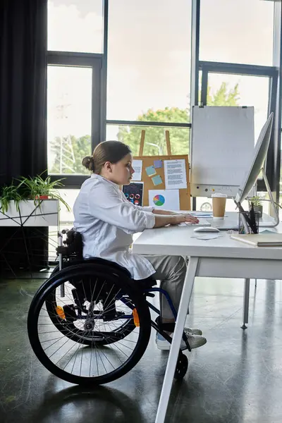 Tekerlekli sandalyedeki genç bir iş kadını modern bir ofis alanında bilgisayarında çalışıyor..