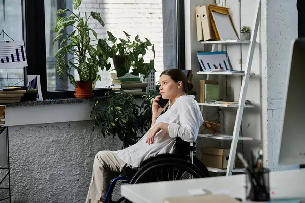 Tekerlekli sandalyedeki genç bir kadın modern bir ofiste çalışırken telefonunu kullanıyor..