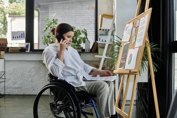 stock image A young businesswoman in a wheelchair sits at a desk in a modern office, working on a project and talking on the phone.