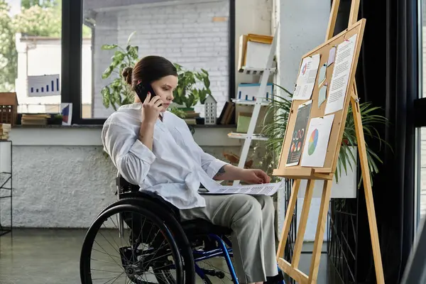 Stock image A young businesswoman in a wheelchair sits at a desk in a modern office, working on a project and talking on the phone.