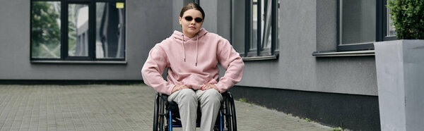 Молодая женщина в розовой толстовке сидит в инвалидном кресле возле здания.