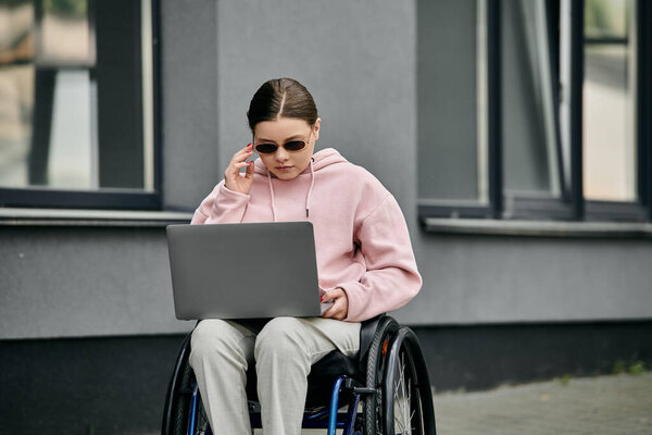 Молодая женщина сидит в инвалидной коляске снаружи, используя ноутбук. На ней розовая толстовка и солнечные очки..