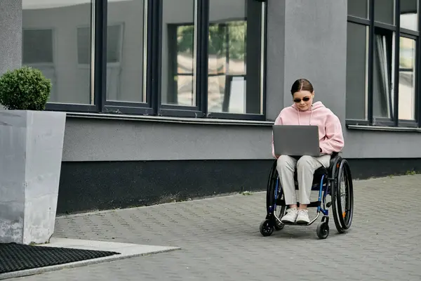 Stock image A young woman in a wheelchair is working on her laptop outside a building. She is wearing a pink hoodie and sunglasses.